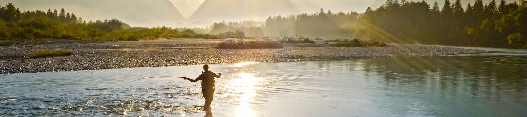 A person is fishing in a river during sunset with mountains and trees in the background, creating a peaceful and scenic landscape.