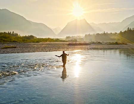 A person is fishing in a river during sunset with mountains and trees in the background, creating a peaceful and scenic landscape.