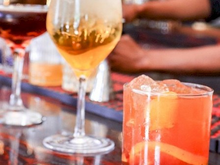 The image shows two cocktails on a table, one with a yellowish drink in a tall glass and another with an orange-red drink in a shorter glass.
