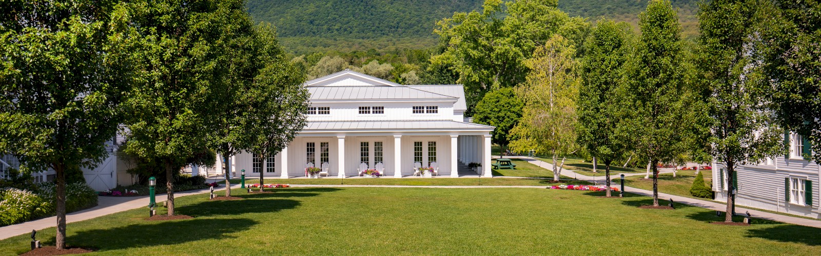 A scenic view of a white building with a porch, surrounded by green trees, colorful flowers, and a mountain backdrop under a blue sky.