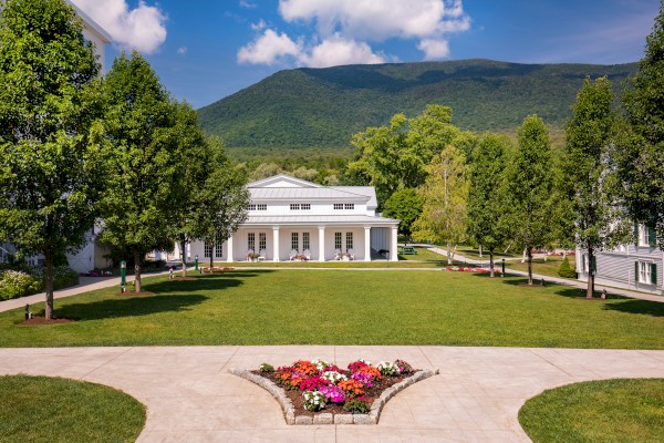 A peaceful garden with a white building, trees, flowers, and a mountain backdrop on a clear, sunny day.