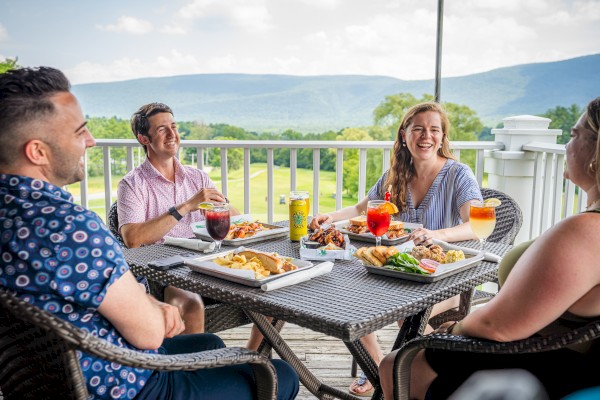 A group of five people enjoying a meal and drinks on a balcony with green fields and mountains in the background.