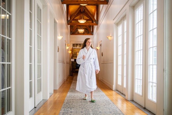 A woman in a white bathrobe and slippers walks down a bright hallway, holding a glass, with sunlight streaming through large windows.