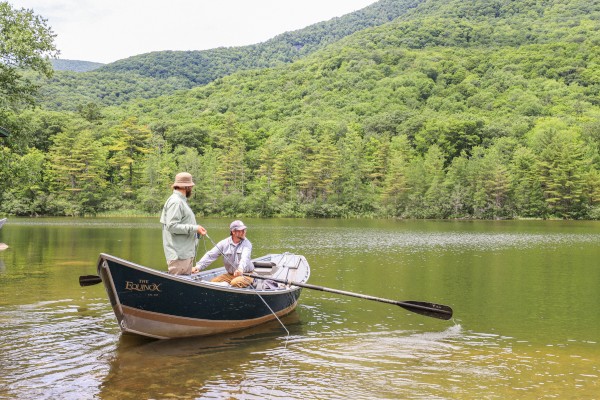 Two men are fishing on a boat in a peaceful, green lake surrounded by lush trees and hills.