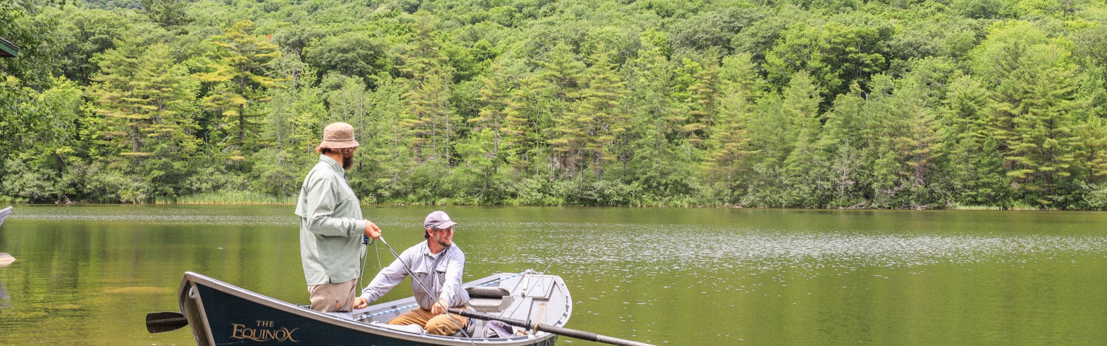 Two men fishing from a boat on a peaceful lake surrounded by lush, green mountains and dense forest.