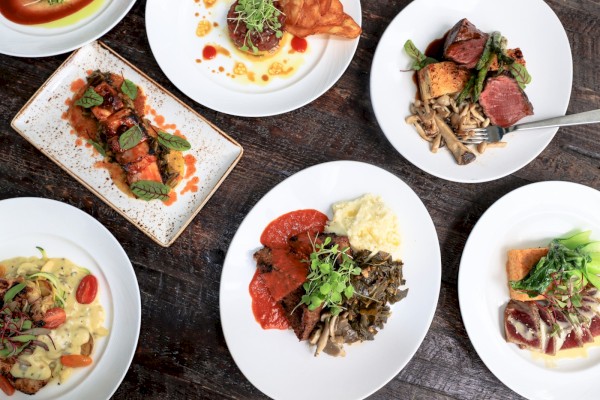 A variety of beautifully plated gourmet dishes, including meats, vegetables, and sauces, arranged on a dark wooden table.