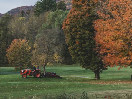 A tractor is mowing a golf course with colorful fall trees and mountains in the background.