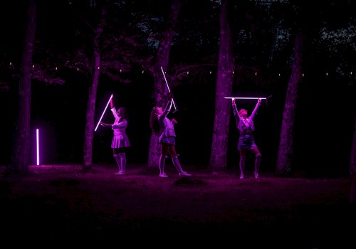 Three women perform with glowing purple rods in a dark, wooded outdoor setting illuminated by neon lights.