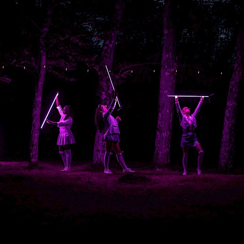 Three women perform with glowing purple rods in a dark, wooded outdoor setting illuminated by neon lights.