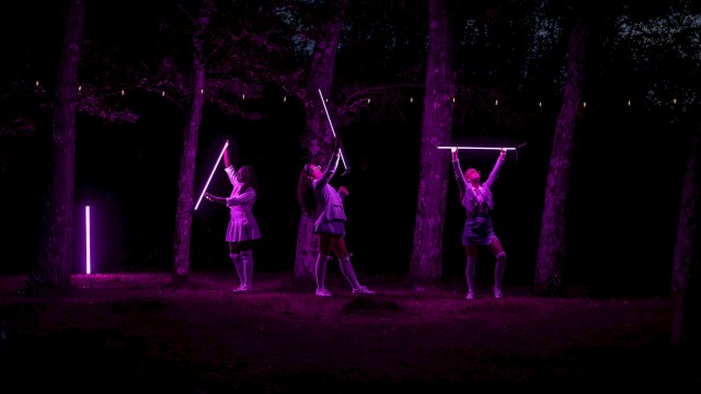 Three women perform with glowing purple rods in a dark, wooded outdoor setting illuminated by neon lights.