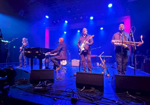A band performs on stage with blue and purple lighting, featuring a pianist, guitarist, bassist, and saxophonist.