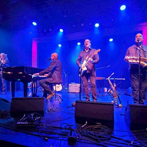 A band performs on stage with blue and purple lighting, featuring a pianist, guitarist, bassist, and saxophonist.