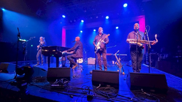 A band performs on stage with blue and purple lighting, featuring a pianist, guitarist, bassist, and saxophonist.