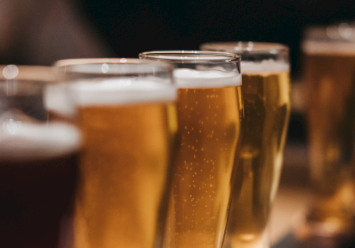 Several glasses of beer lined up on a table, likely at a social gathering or pub, with a warm, blurry background.