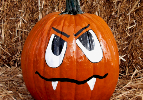 A painted pumpkin with an angry face, featuring large eyes, frowning eyebrows, and two small fangs sitting on straw.