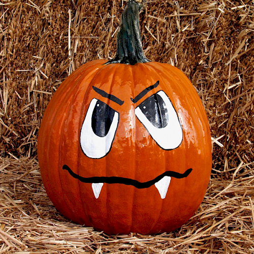 A painted pumpkin with an angry face, featuring large eyes, frowning eyebrows, and two small fangs sitting on straw.