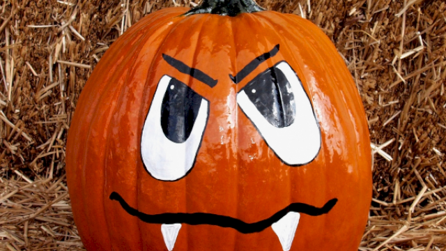 A painted pumpkin with an angry face, featuring large eyes, frowning eyebrows, and two small fangs sitting on straw.