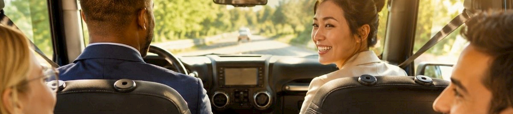 A group of five diverse professionals is happily chatting and smiling during a car ride on a sunny day.