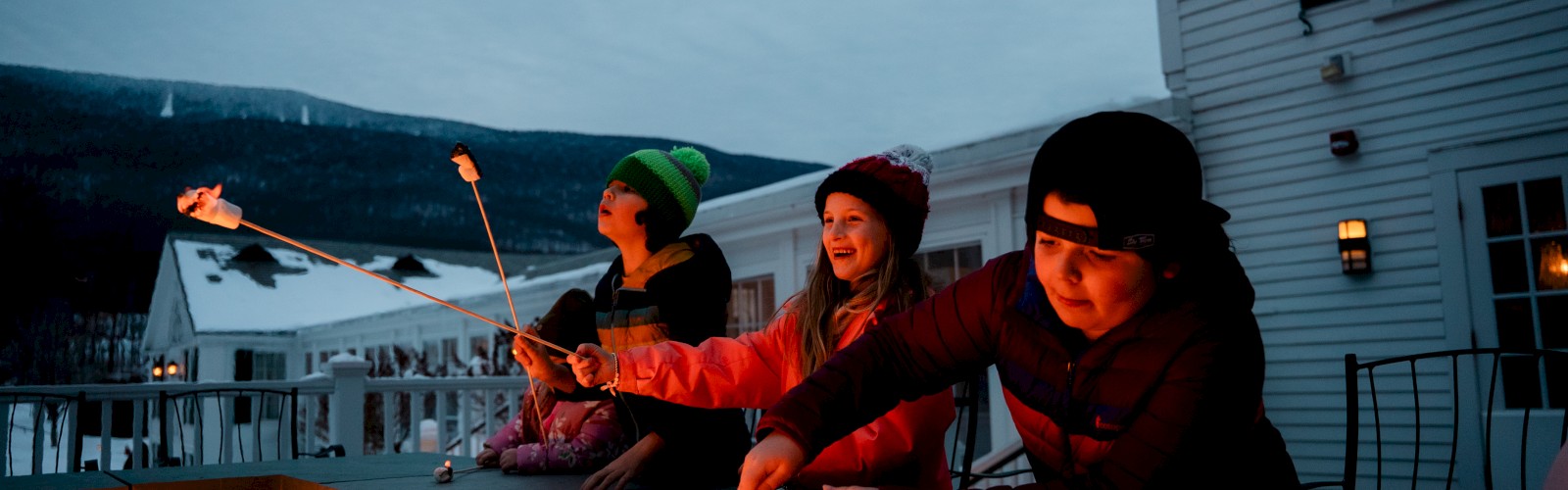 Kids roasting marshmallows around a fire pit on a snowy evening, enjoying a cozy outdoor gathering at a house.