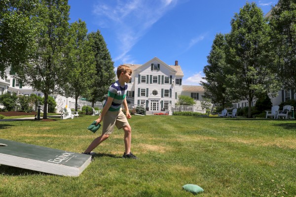 A boy throws a bean bag on a sunny lawn in front of a large white house with trees and benches.