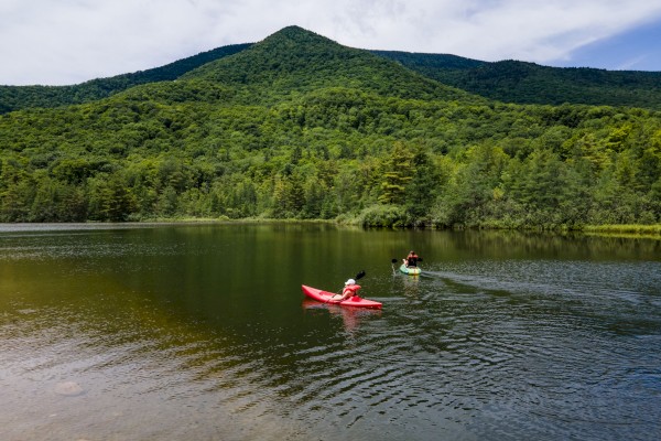 Two people are kayaking on a calm lake with a lush green mountainside and partly cloudy sky in the background.