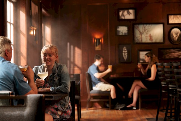 People are enjoying drinks and conversations in a cozy, warmly-lit bar with dark wooden walls and framed artwork.
