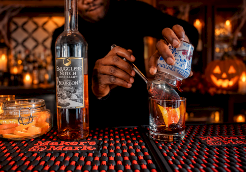 A bartender pours a drink from a bottle of bourbon at a Halloween-themed bar with a glowing pumpkin in the background.