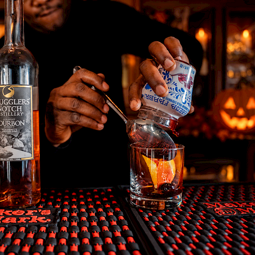 A bartender pours a drink from a bottle of bourbon at a Halloween-themed bar with a glowing pumpkin in the background.