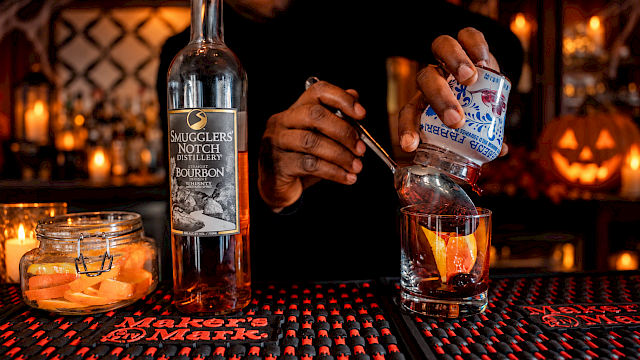 A bartender pours a drink from a bottle of bourbon at a Halloween-themed bar with a glowing pumpkin in the background.