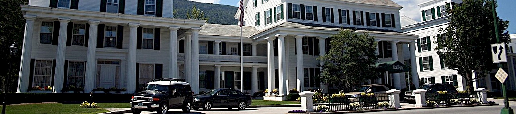 A large, white, multi-story building with columns, parked cars, trees, and a clear blue sky with clouds.