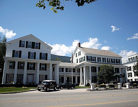 A large, white, multi-story building with columns, parked cars, trees, and a clear blue sky with clouds.