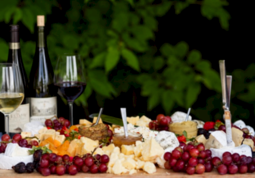 A cheese and fruit platter with wine, featuring grapes, cheeses, and bottles of wine on a wooden table outdoors.