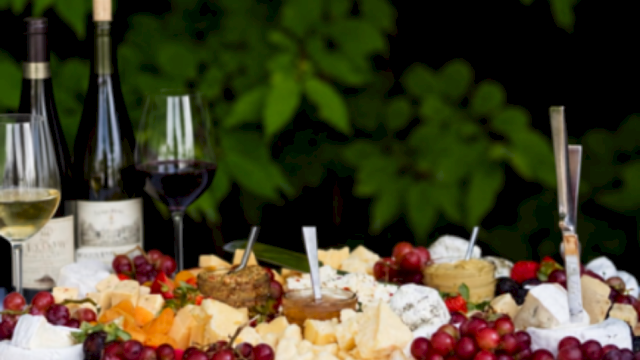 A cheese and fruit platter with wine, featuring grapes, cheeses, and bottles of wine on a wooden table outdoors.