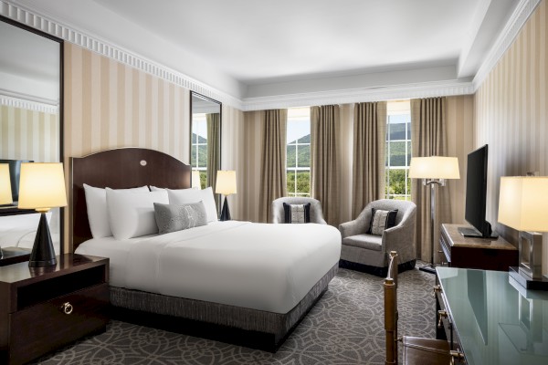 A hotel room with a large bed, lamps, armchairs, a TV, and windows showing a mountain view.