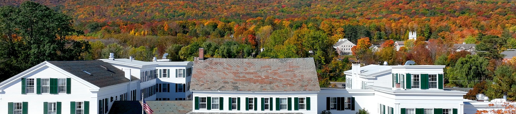 A large white hotel building with green shutters is set against a colorful autumn mountain landscape with trees in orange, red, and yellow.