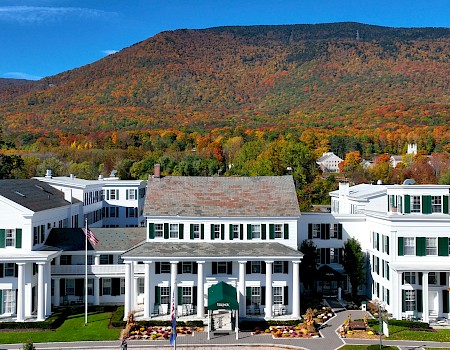 A large white hotel building with green shutters is set against a colorful autumn mountain landscape with trees in orange, red, and yellow.