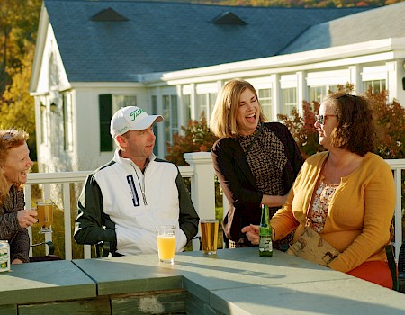 Four friends are enjoying drinks and laughing together outdoors on a sunny day at a white house with fall foliage.