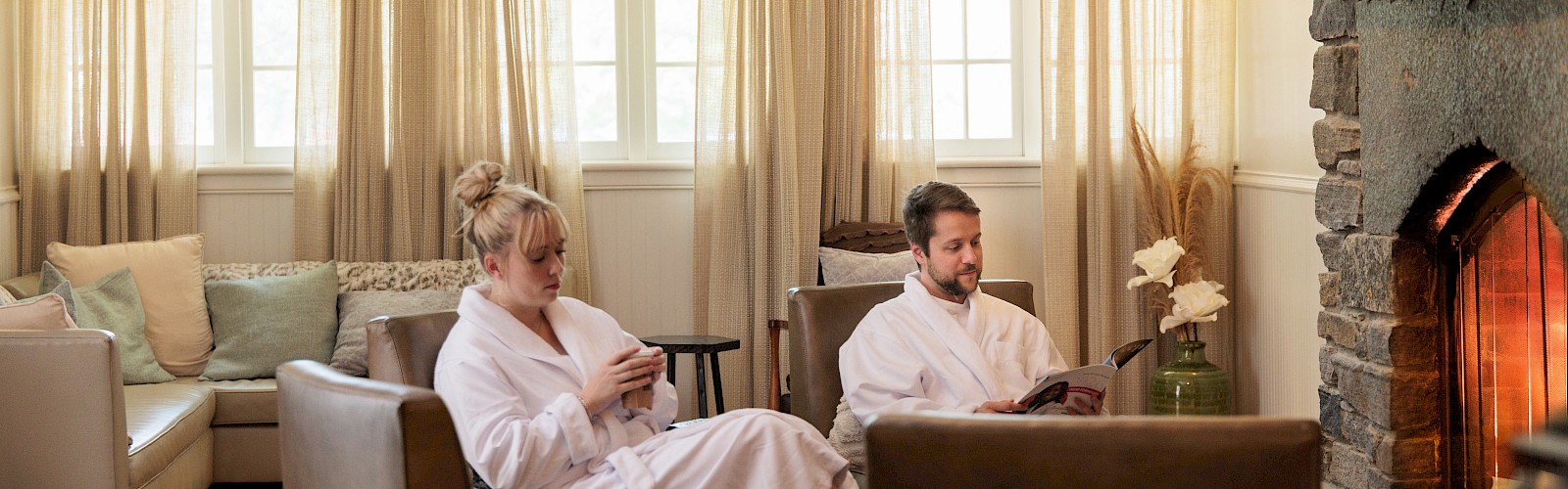 A man and a woman in white robes relax by a fireplace, one reading a magazine and the other on her phone in a cozy living room.