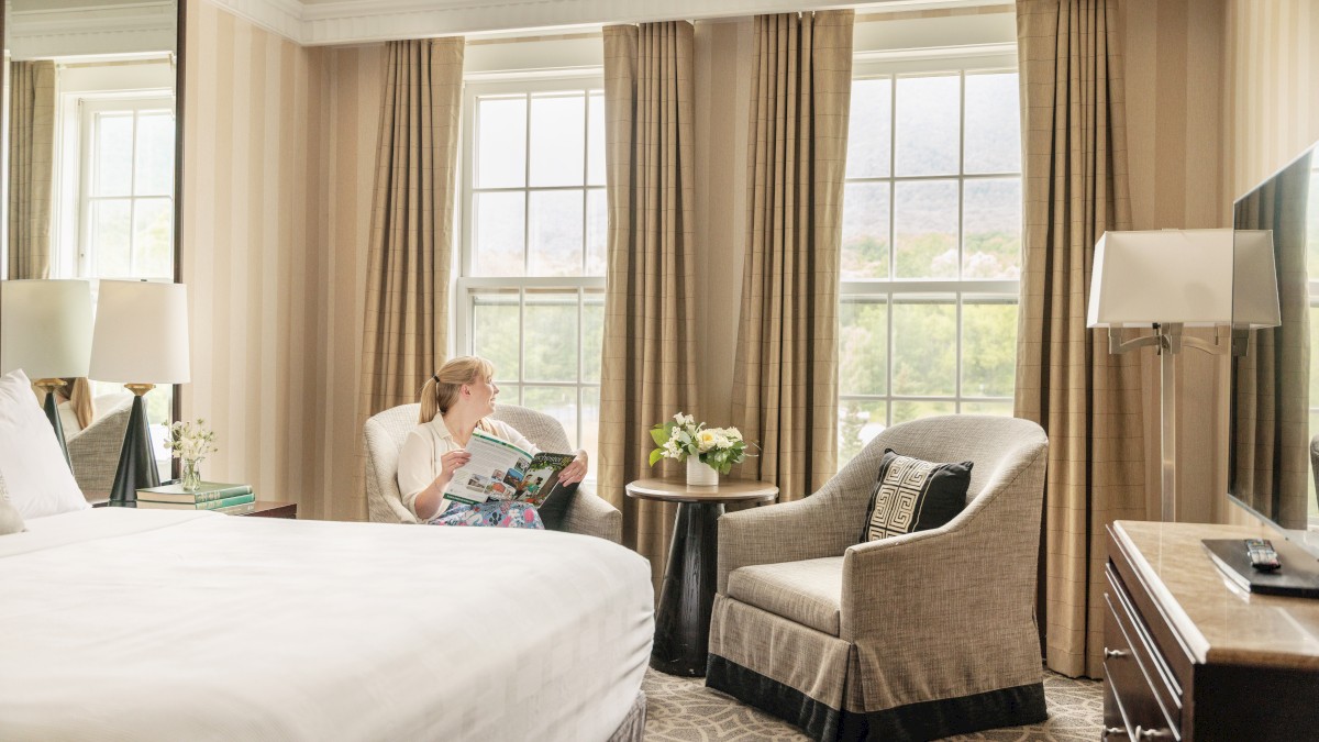 A woman sits by a large window in a hotel room, reading a magazine while relaxing on a beige armchair amidst cozy decor.
