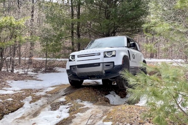 An electric off-road vehicle is navigating a snowy, wooded trail with uneven terrain in this outdoor scene.