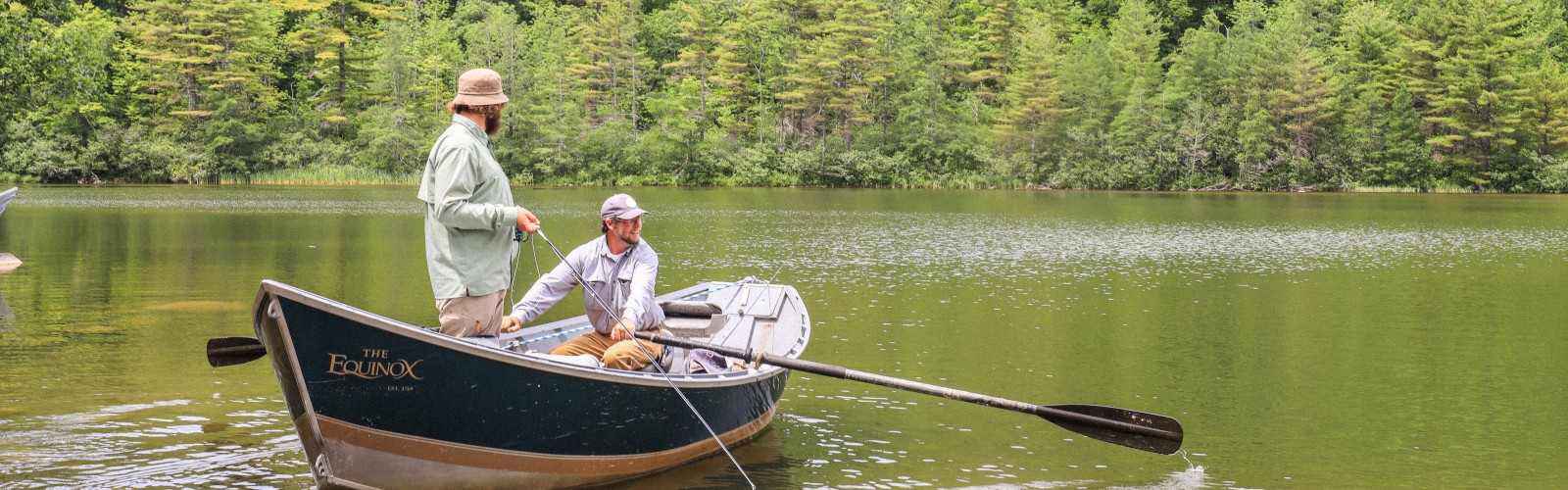 Two people are in a boat on a calm lake surrounded by lush, green hills. One is standing, and the other is seated, with fishing rods visible in the boat.