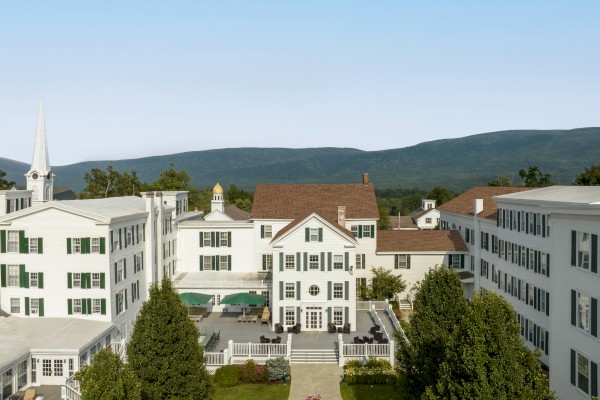 A large white colonial-style building with several wings, surrounded by greenery and featuring mountains in the background under a clear blue sky.