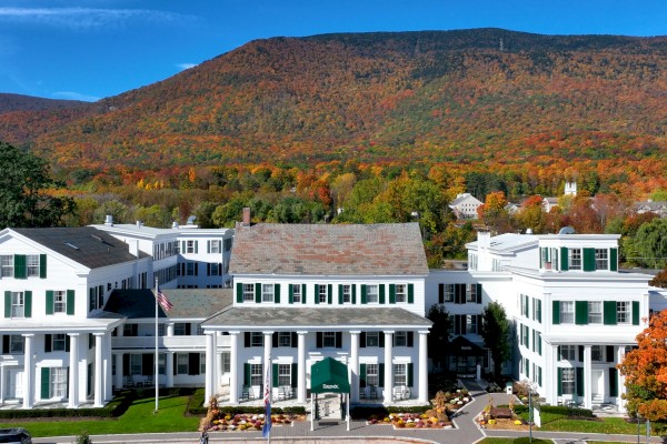 A large white building with green shutters is set against a backdrop of colorful autumn trees and a mountain under a clear blue sky.