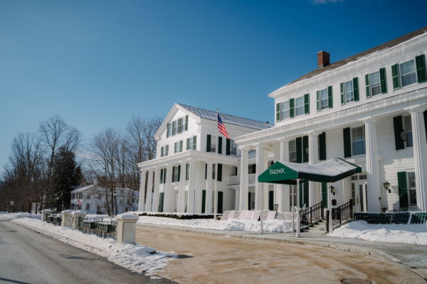 A snowy street scene with large white buildings featuring green shutters and an American flag, under a clear blue sky.