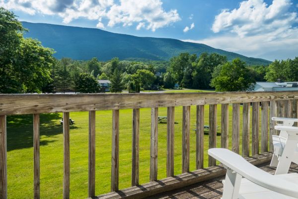 A wooden deck with white chairs overlooking a green lawn, trees, and a mountain under a blue sky with clouds.