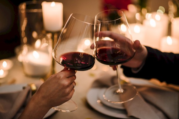 Two people clinking red wine glasses in a cozy, candle-lit setting, suggesting a romantic or celebratory atmosphere.