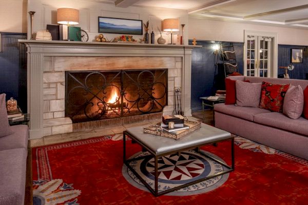 This image shows a cozy living room with a lit fireplace, sofas, a red rug, a coffee table, and various decorative items on the mantle.