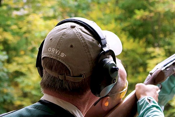 A person wearing protective headphones and a cap takes aim with a shotgun in an outdoor wooded area.