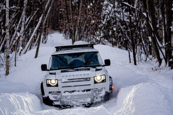 A white SUV drives through a snowy forest path, surrounded by snow-covered trees and deep snow on the ground.