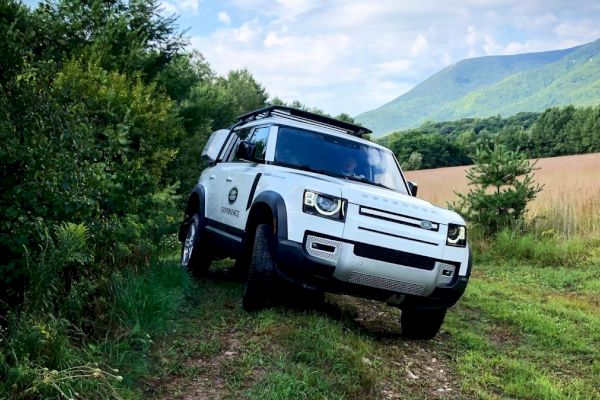A white Land Rover off-road vehicle is parked on a grassy path surrounded by nature, with mountains visible in the background.